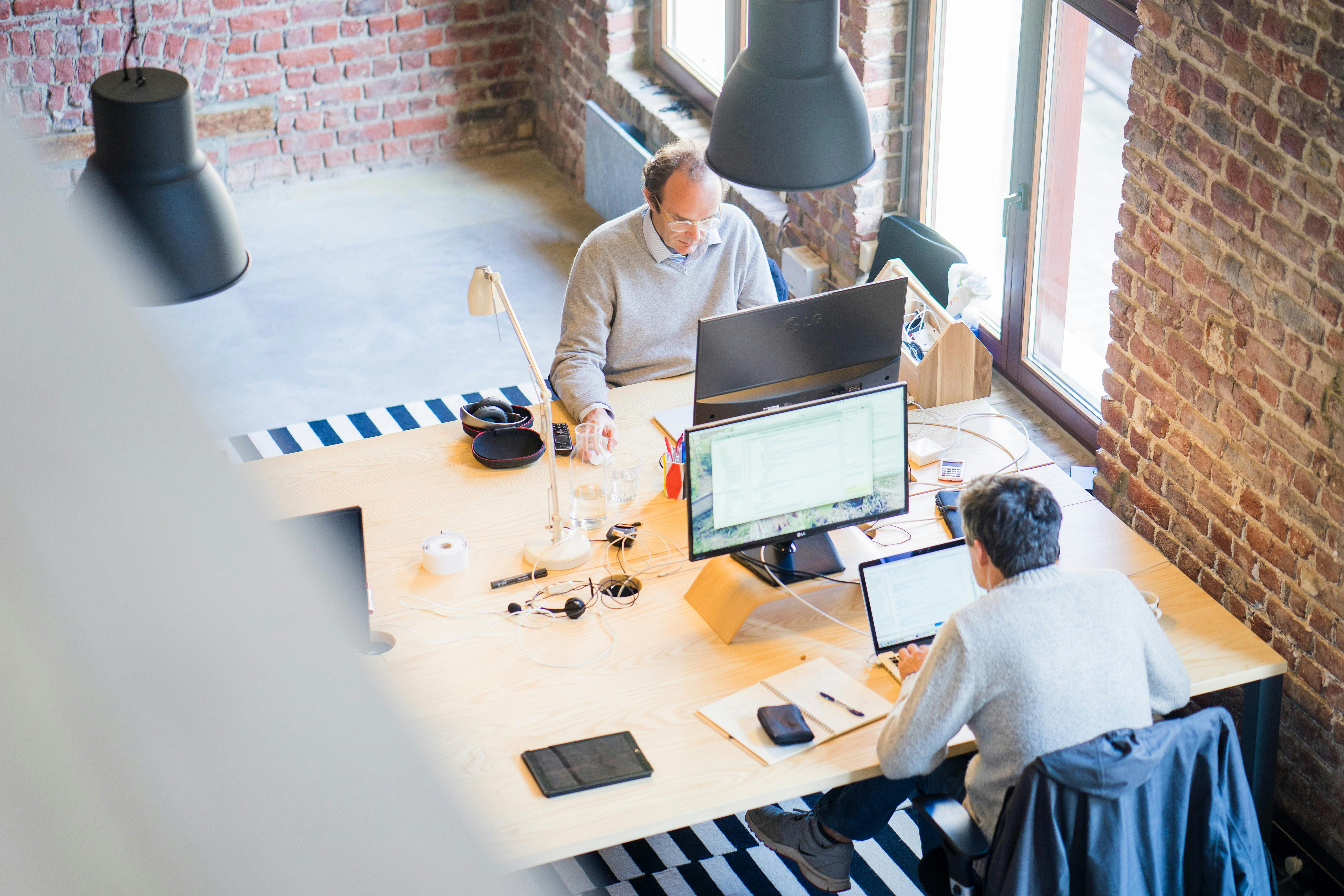 Two professionals working in a modern open office space with wooden desks, computers, and exposed brick walls, creating a productive and collaborative work environment. They are working on a approach on how to create a website.